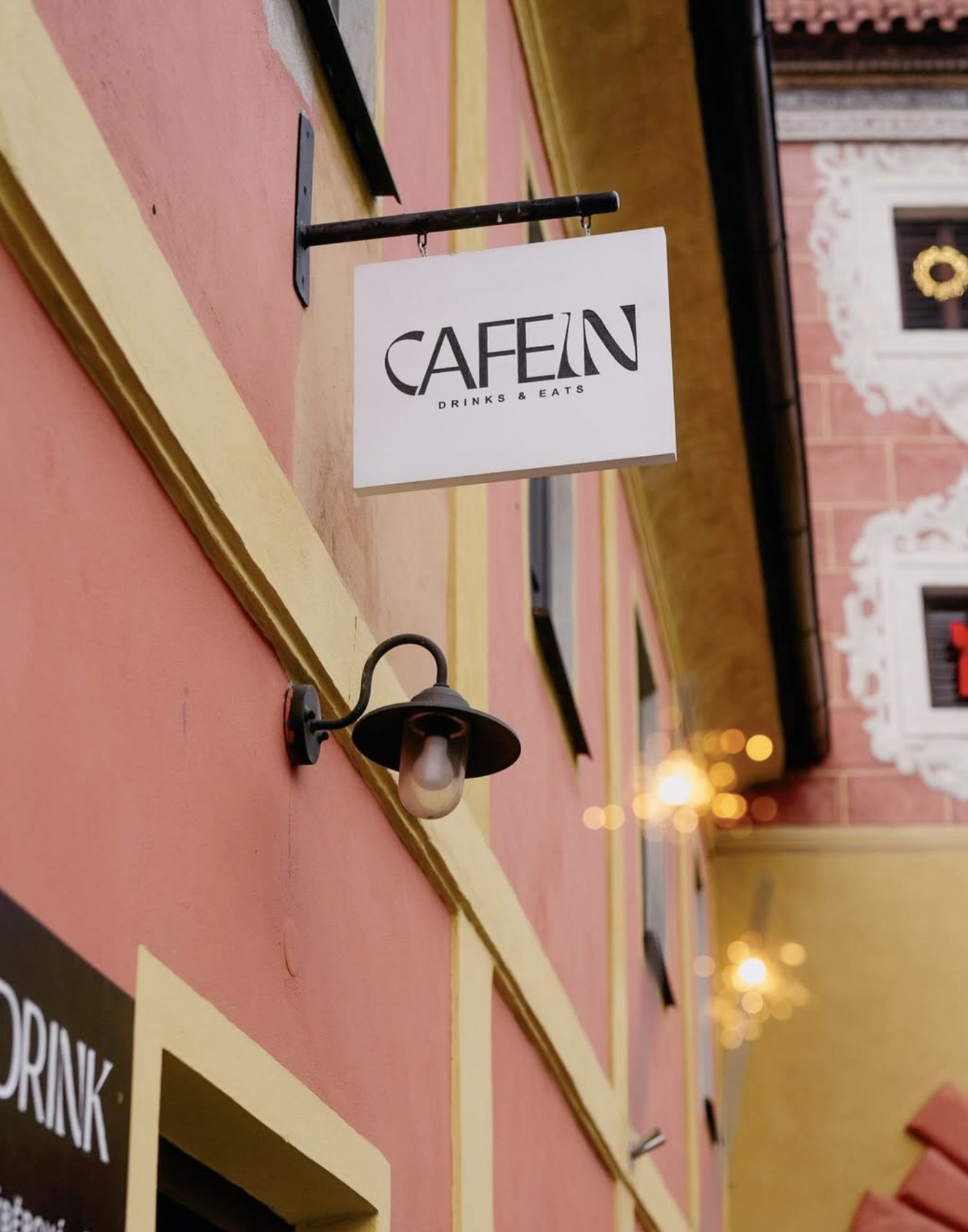 The CAFEIN sign hanging above a pink and yellow facade, with a lantern style wall lamp below it